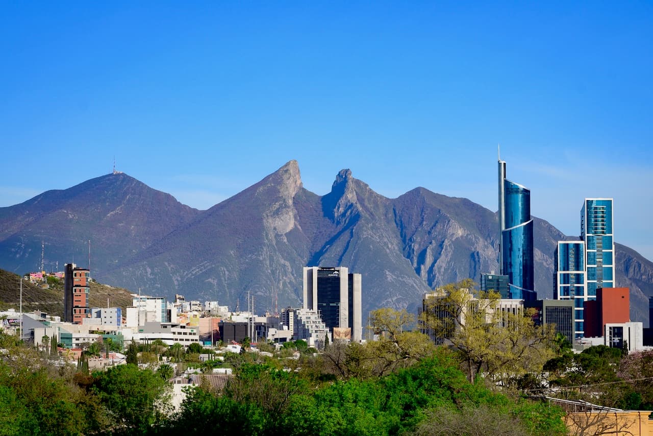 Vista de una ciudad con edificios modernos y altos en primer plano, rodeada de vegetación, y al fondo se destacan montañas con picos afilados bajo un cielo azul claro.