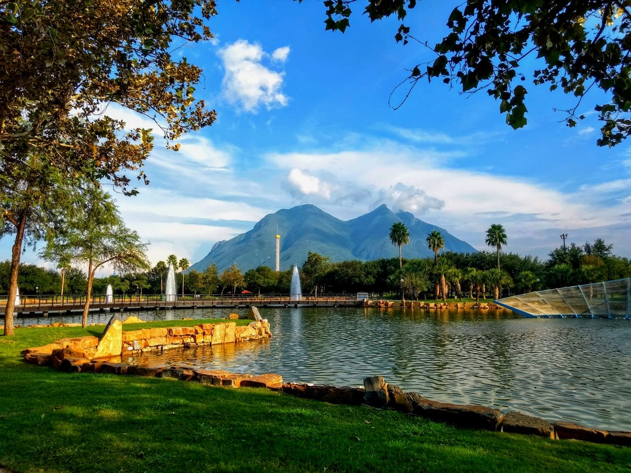 Un parque junto a un lago con fuentes que brotan agua, rodeado de árboles y palmeras. Al fondo se pueden ver montañas bajo un cielo parcialmente nublado. Hay un camino de madera que se extiende a lo largo del lago.