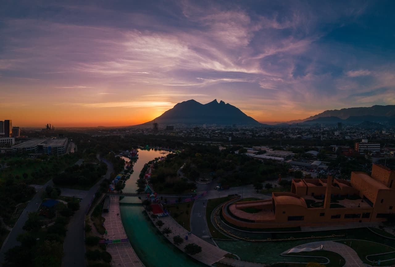 Un paisaje de Monterrey al atardecer, con la imponente montaña del Cerro de la Silla al fondo. Se observa un río serpenteante y áreas verdes, así como edificios y una estructura arquitectónica moderna en primer plano. El cielo muestra tonalidades de naranja y púrpura, creando un ambiente cálido y sereno.