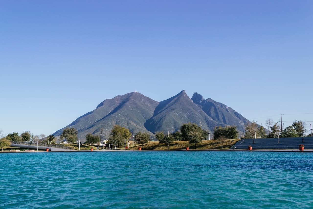 Un paisaje con un lago de aguas azuladas en primer plano, y al fondo se destacan montañas imponentes bajo un cielo despejado. Hay vegetación a los lados y estructuras en la orilla del lago.