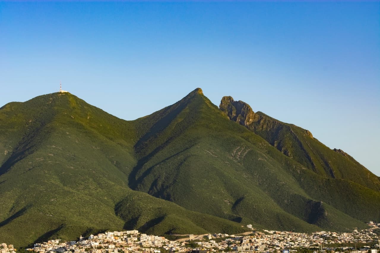 Una vista de montañas verdes bajo un cielo claro, con un pico prominente que tiene una antena en la cima y otra formación rocosa en la parte derecha. Al pie de las montañas se ve una ciudad con varias casas.