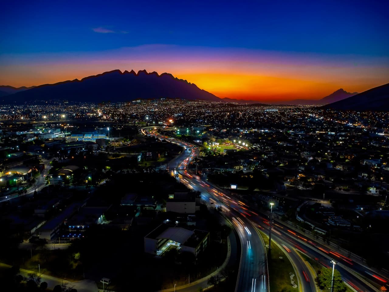 Vista nocturna de una ciudad iluminada, con un gran horizonte montañoso al fondo. En primer plano, se observa una carretera con luces de vehículos en movimiento y una serie de edificios dispersos en un área urbana. El cielo presenta un colorido degradado que va desde el naranja hacia el azul oscuro, sugiriendo el ocaso.