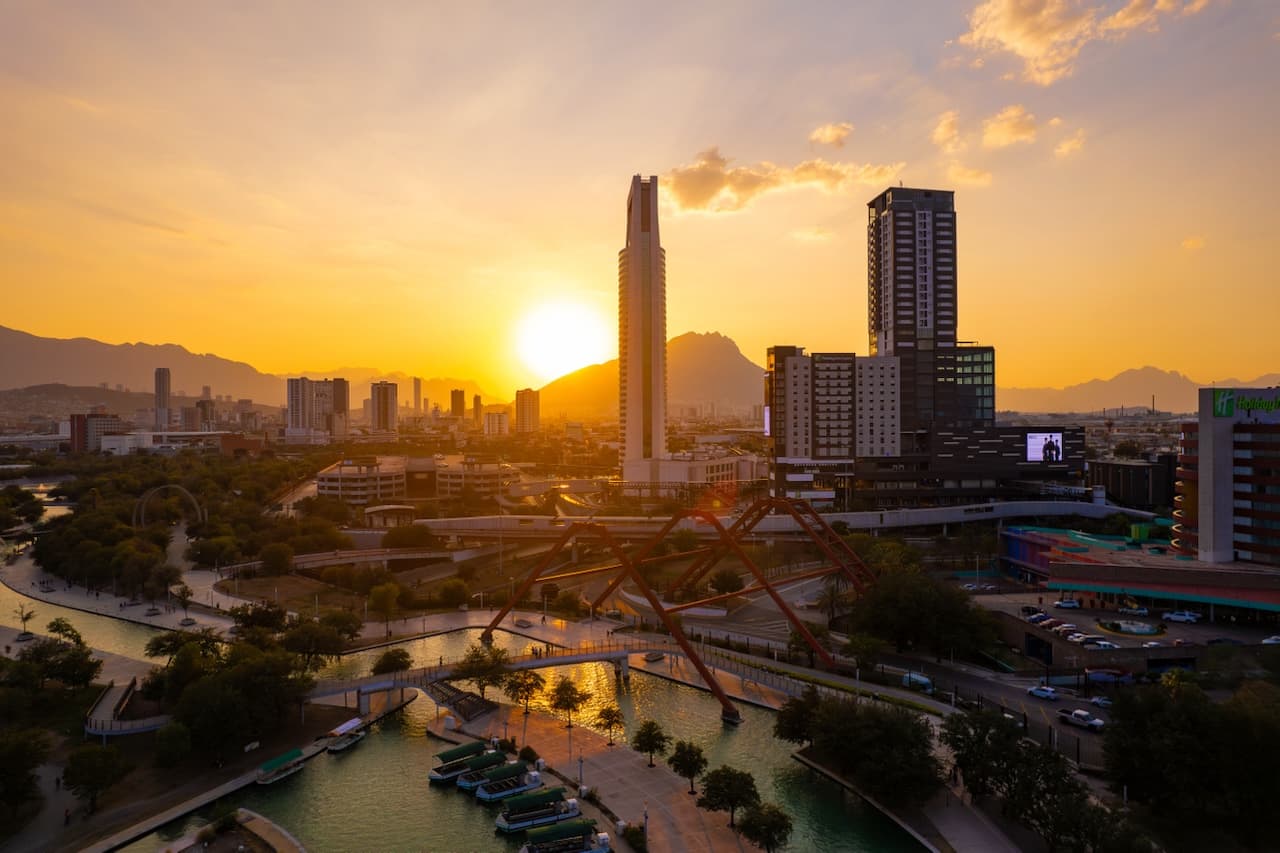 Vista panorámica de una ciudad al atardecer, con un sol amarillo cerca del horizonte. Se destacan edificios modernos y una montaña al fondo, acompañados de un canal y áreas verdes. En el primer plano, se pueden ver embarcaciones en el agua y un acceso peatonal que conecta las distintas áreas. La atmósfera es cálida y tranquila.