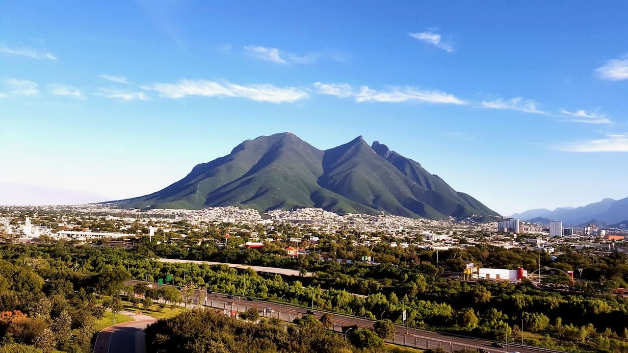 Vista panorámica de montañas con forma pronunciada, rodeadas de áreas urbanas y vegetación. El cielo es azul con algunas nubes dispersas. Se observan edificios y carreteras en el primer plano, junto a árboles y áreas verdes.