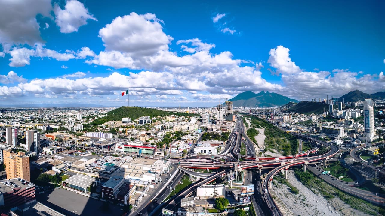 Vista panorámica de una ciudad, con edificios altos, calles y avenidas que se entrelazan. En el fondo, se observa una montaña, mientras que el cielo está despejado con algunas nubes. También se destaca una gran bandera de México en una colina.