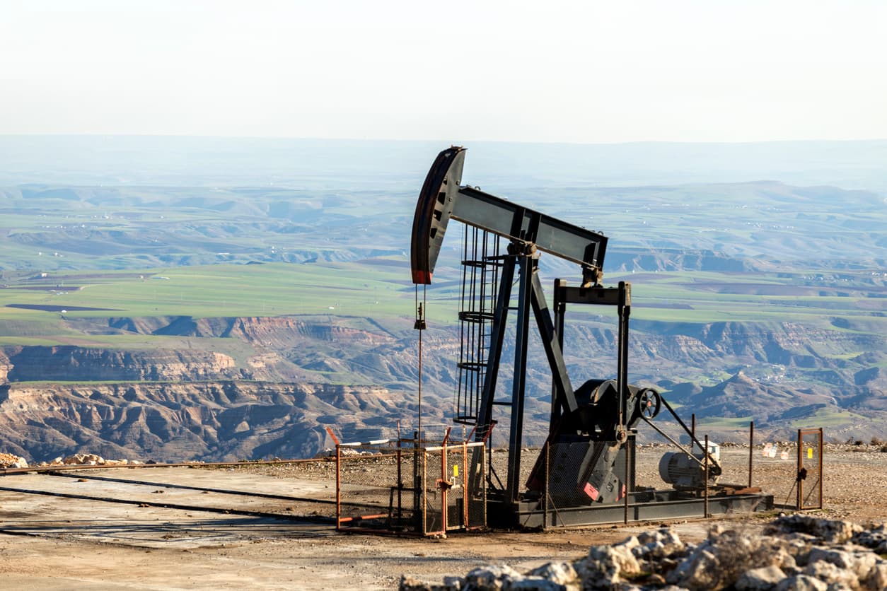 Una bomba de extracción de petróleo se encuentra en una área montañosa, con un paisaje que muestra colinas verdes y formaciones rocosas en el fondo. La instalación parece estar bien cuidada, y la escena refleja un entorno industrial en medio de la naturaleza.