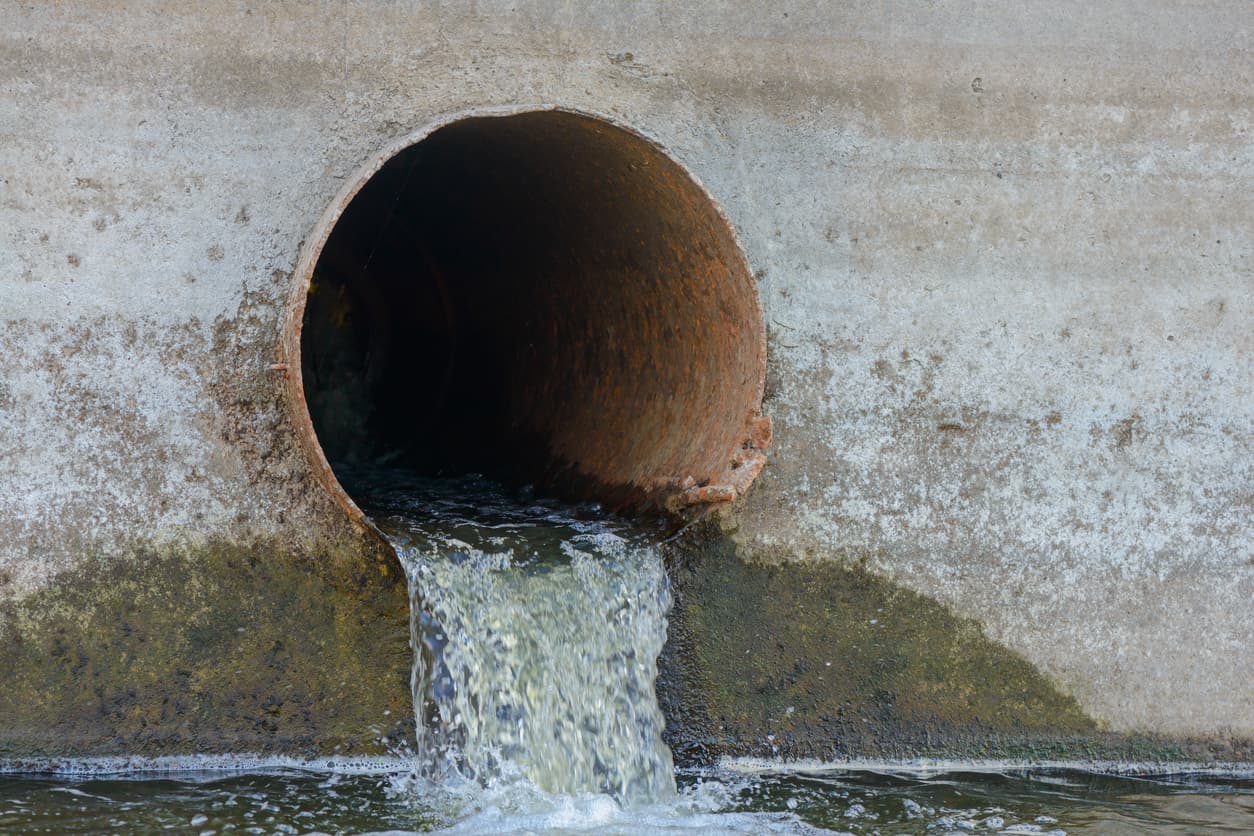 Se observa un tubo helical de concreto donde sale agua, creando una corriente que fluye hacia un cuerpo de agua cercano. La zona alrededor del tubo muestra manchas de humedad y suciedad.