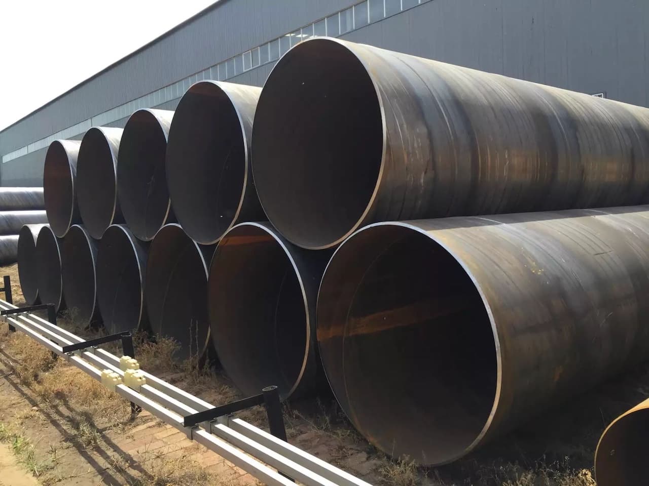 Se observa una serie de tubos helicoidales apilados, de gran diámetro, con un acabado metálico y aspecto rústico. Al fondo, se puede ver un edificio industrial de estructura metálica y ventanales. También hay tubos más delgados dispuestos horizontalmente en la parte inferior de la imagen. La escena tiene un ambiente exterior con vegetación seca alrededor.